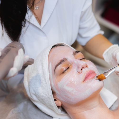 Beautician with a brush applies a white moisturizing mask to the face of a young girl client in a spa beauty salon