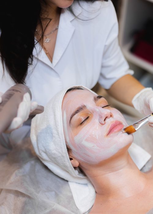 Beautician with a brush applies a white moisturizing mask to the face of a young girl client in a spa beauty salon